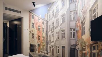 A hotel room with a mural of a European street, a bed, a desk, and a TV.
