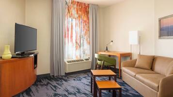 Ein Hotelzimmer mit einem Flachbildfernseher auf einer Holzkommode, einem grünen Sessel, einem beigen Sofa und einem kleinen Holztisch.