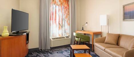 A hotel room with a flat-screen TV on a wooden cabinet, a green chair, a beige sofa, and a small wooden table.