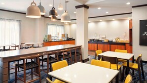 A hotel dining area with multiple tables and chairs, a kitchen in the background, and pendant lighting. - Fairfield Inn & Suites by Marriott Selma Kingsburg (Kingsburg)