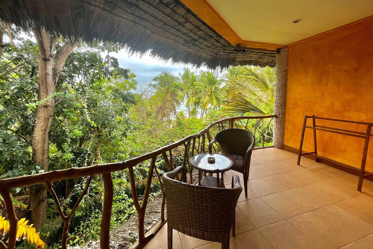 A balcony with wicker chairs and a small table, overlooking a lush garden and palm trees.
