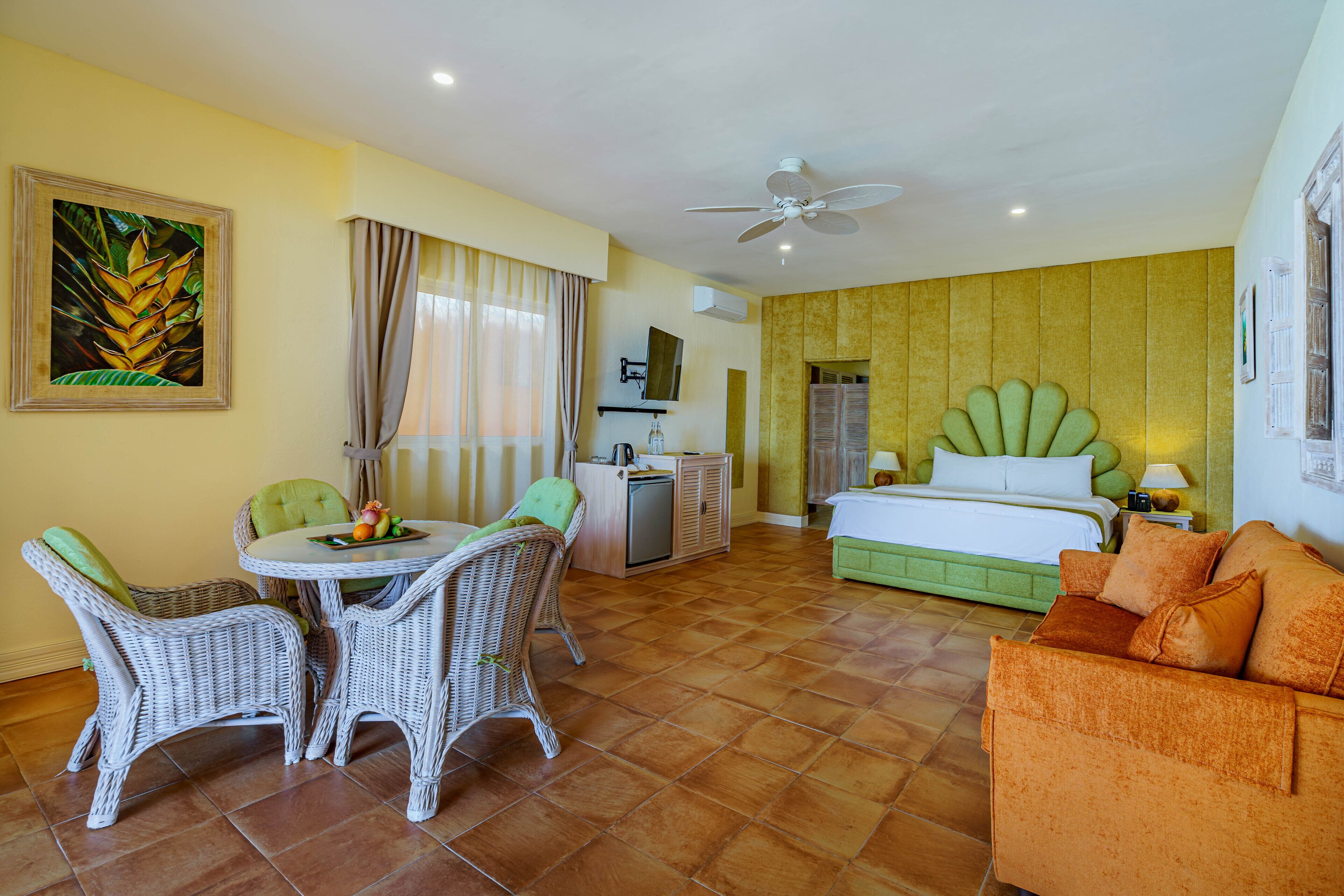 A hotel room with a bed, a green sofa, a dining table with chairs, a TV, and a ceiling fan.