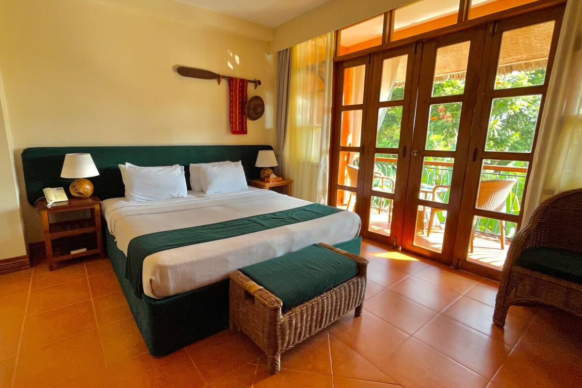 A hotel room with a large bed, two bedside tables with lamps, a wicker chair, and a view of the outdoors through a glass door.