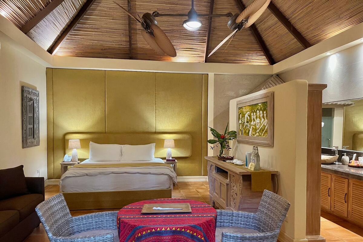 A hotel room with a bed, two chairs, a vanity with a mirror, and a ceiling fan.