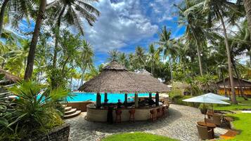 A thatched-roof bar area with a pool view, surrounded by palm trees and a clear sky.