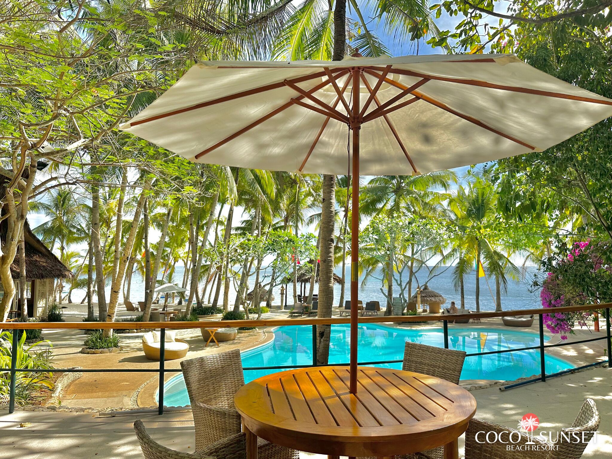 A beach resort with a pool, lounge chairs, and a wooden table under a canopy.