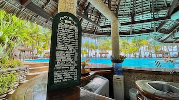 Eine Bar mit einer Speisekarte, die Cocktails und Preise auflistet, Blick auf einen Pool und einer tropischen Umgebung mit Palmen.