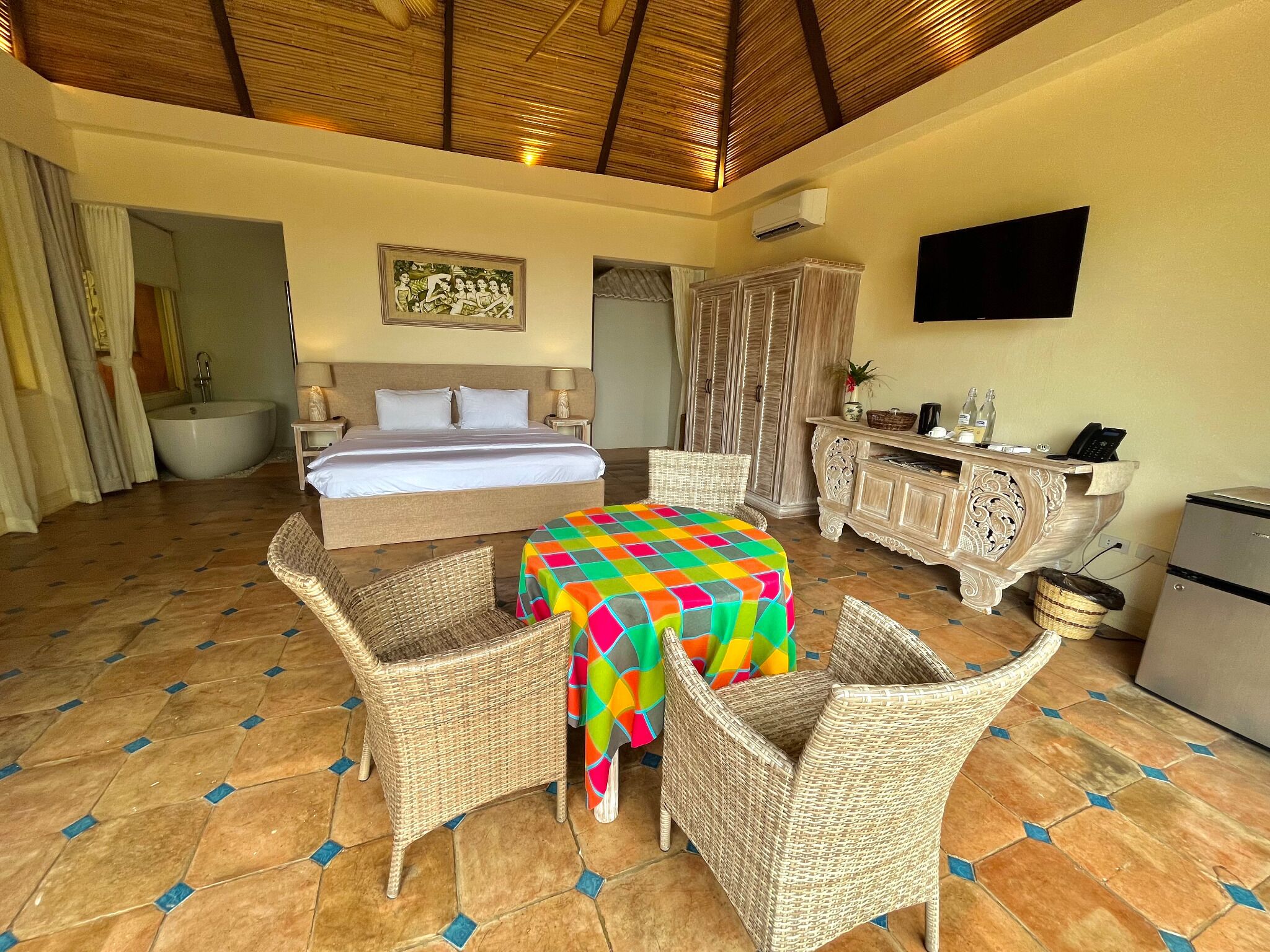 A hotel room with a bed, a dining table with chairs, a television, and a minibar.