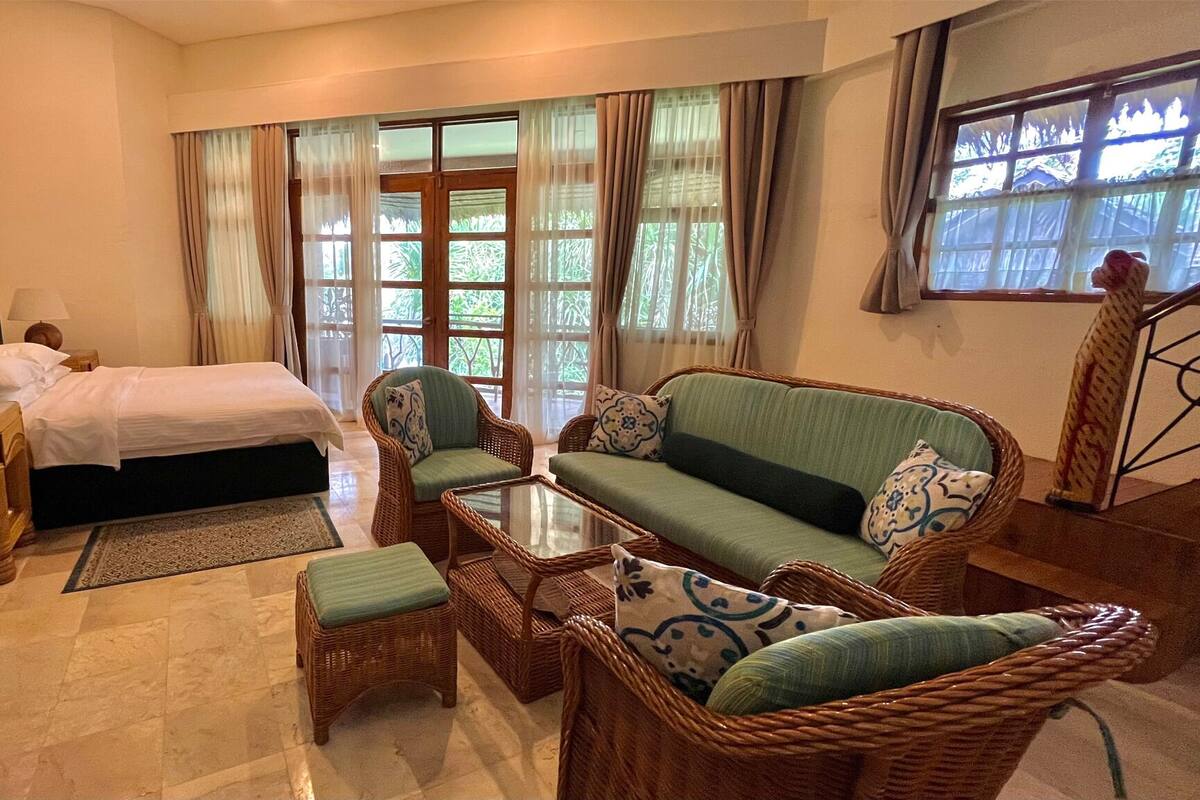A hotel room with a bed, a green sofa, wicker furniture, and a view of greenery through the windows.