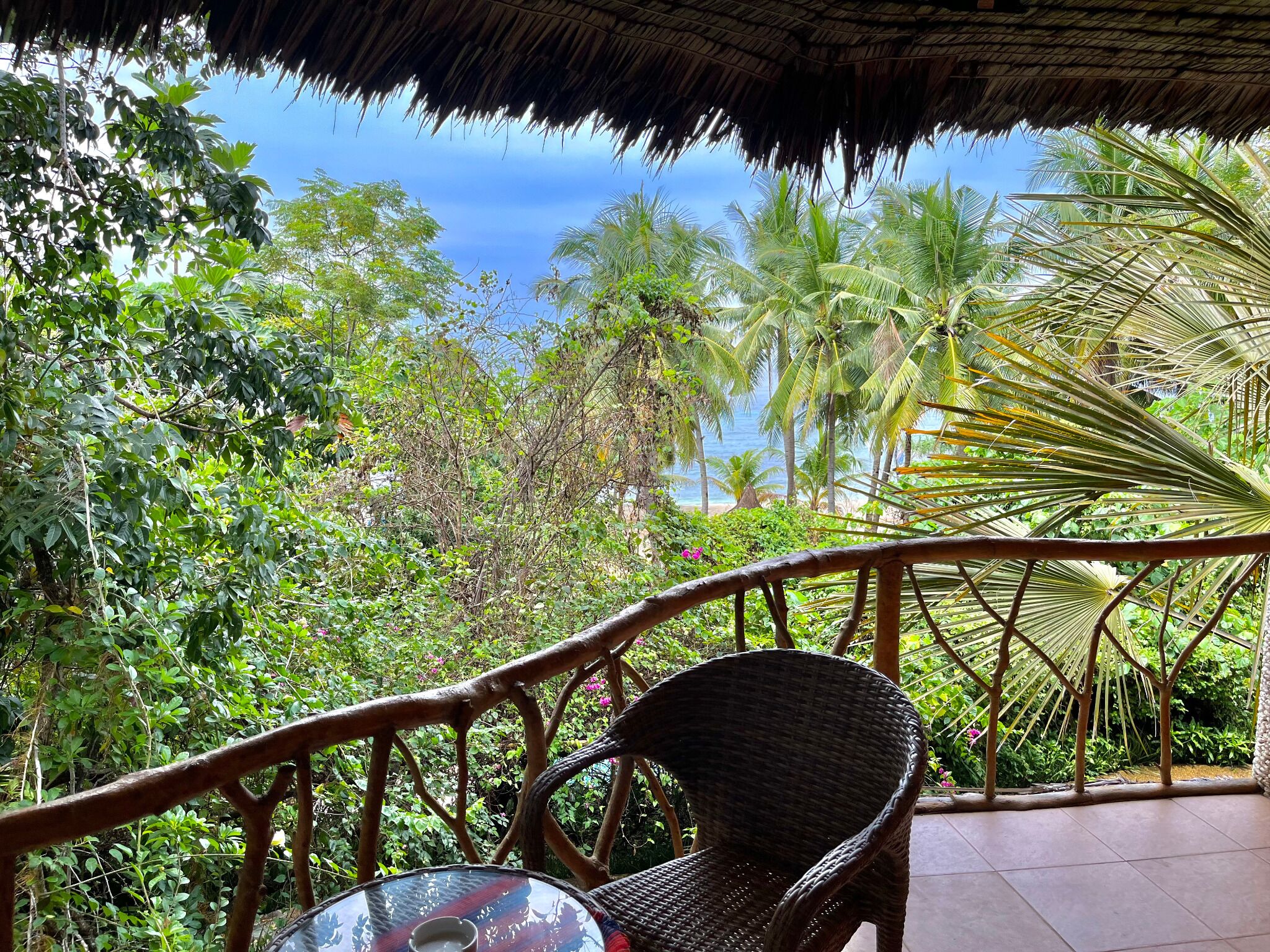 A wicker chair and a glass table on a balcony with a thatched roof, overlooking a lush garden and a beach.