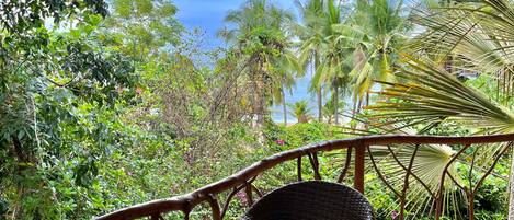 A wicker chair and a glass table on a balcony with a thatched roof, overlooking a lush garden and a beach.
