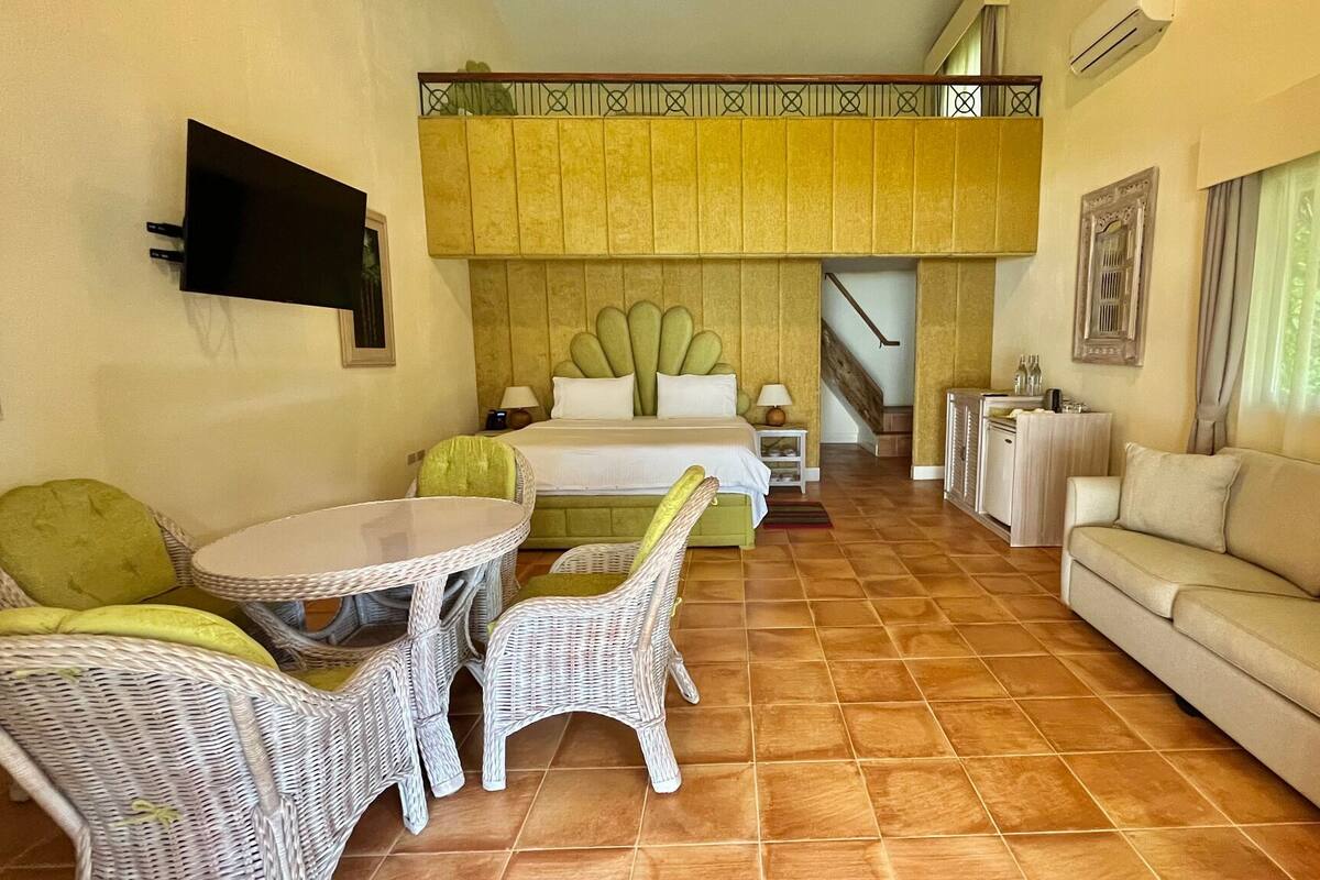 A hotel room with a bed, a sofa, a dining table with chairs, and a TV mounted on the wall.