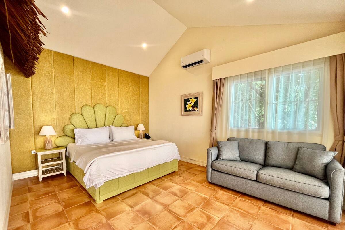 A bedroom with a bed, a grey sofa, a nightstand, an air conditioner, a window with curtains, and a wall with a framed picture.