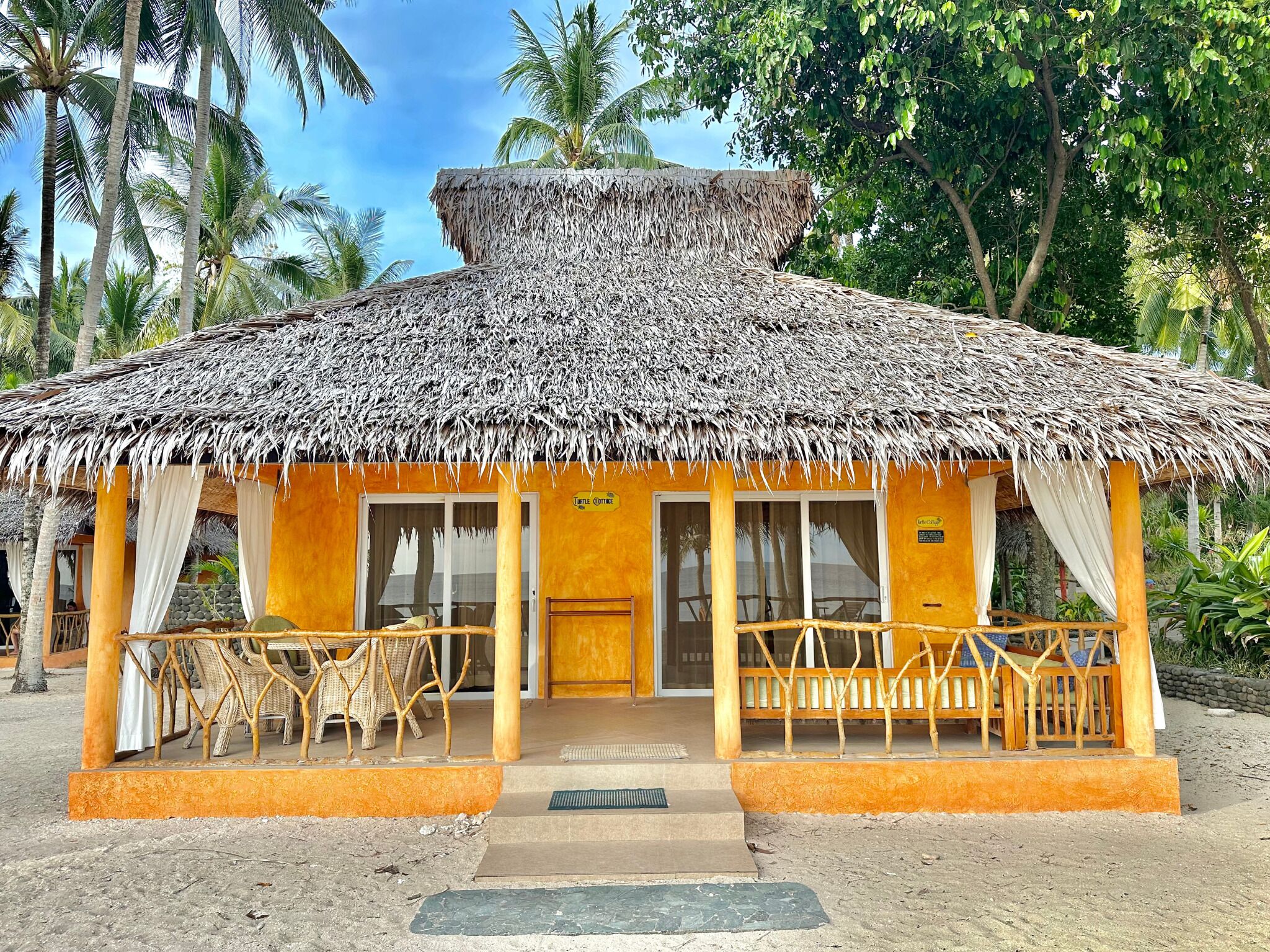 A thatched-roof hut with a porch, wicker furniture, and a sign indicating it is a 'Lounge Cottage'.