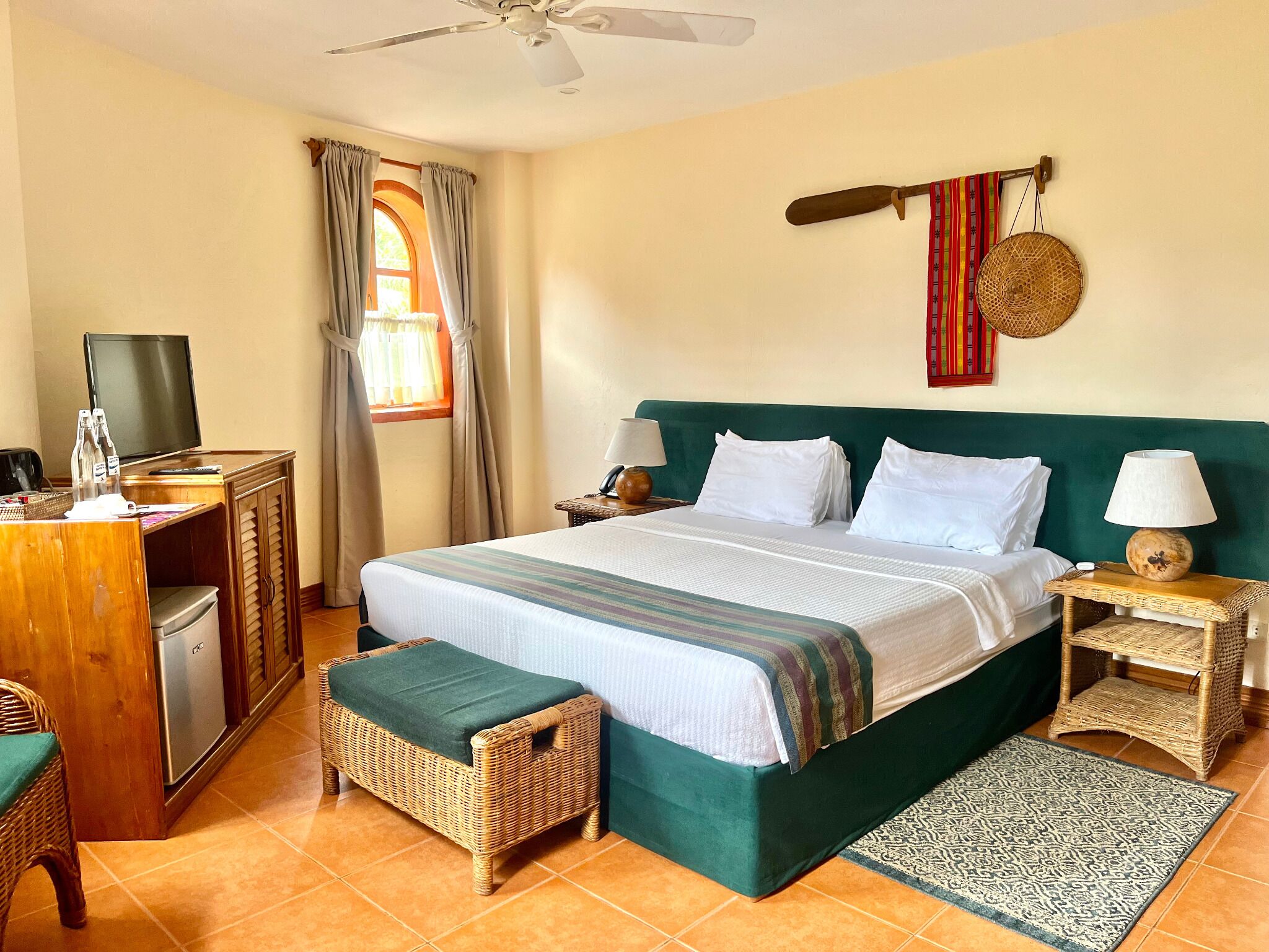 A hotel room with a bed, a desk with a television, a chair, a small refrigerator, and a ceiling fan.