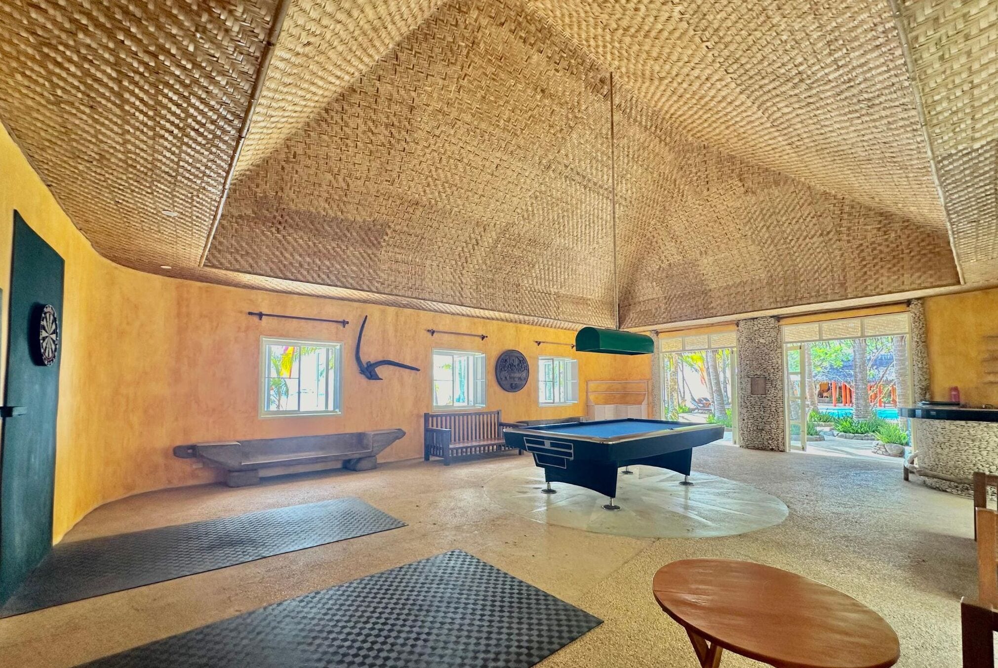 A pool table in a room with a high, woven ceiling and large windows.