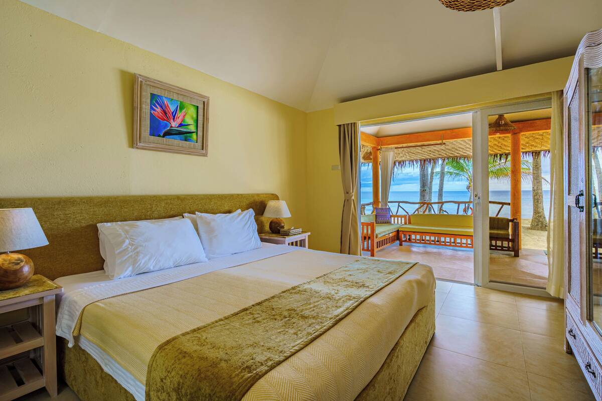 A hotel room with a bed, a nightstand, a lamp, a painting, and a view of the beach through an open door.