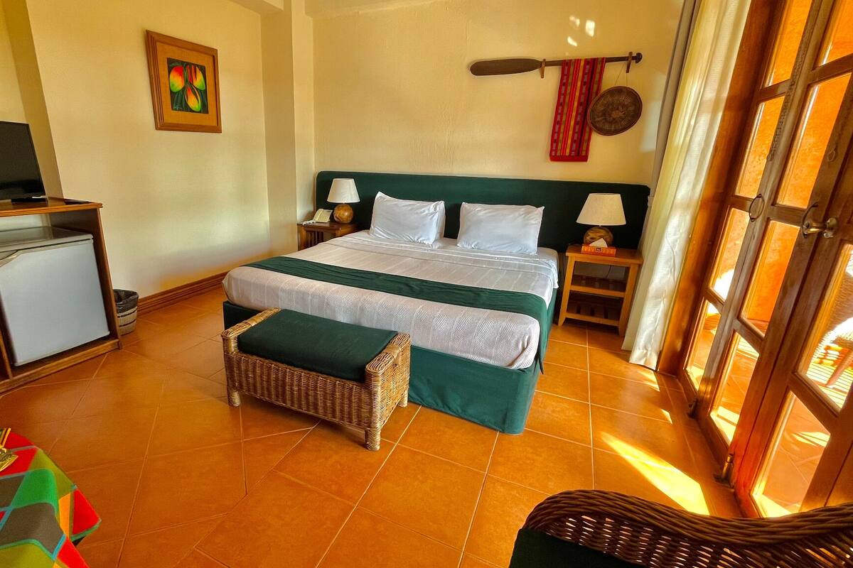 A hotel room with a bed, two bedside tables, a TV, a small fridge, and a wicker chair.