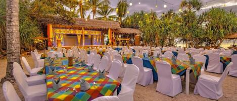 Un espacio para eventos al aire libre con mesas y sillas, un escenario y palmeras.