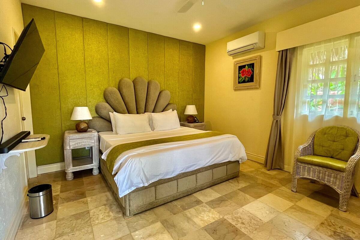 A hotel room with a bed, a television mounted on the wall, a nightstand with a lamp, a chair, and a window with curtains.