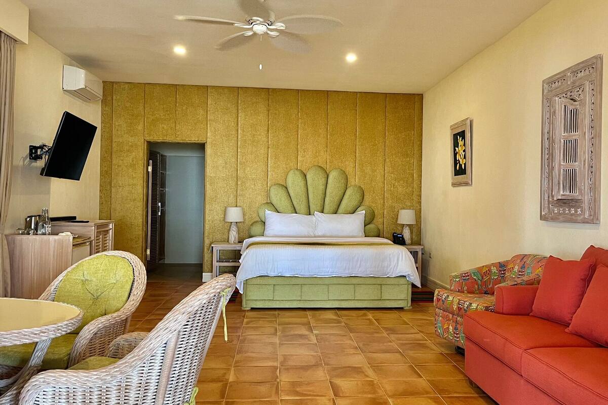 A hotel room with a bed, a television, a ceiling fan, a red sofa, wicker chairs, and a tiled floor.