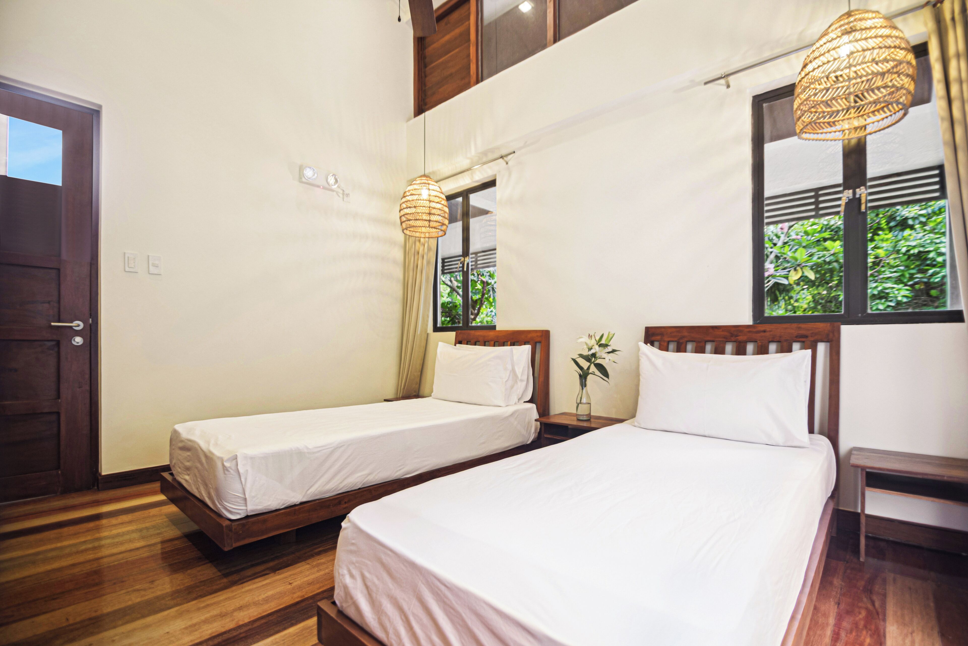A hotel room with two single beds, wooden flooring, a wooden door, and a window with a view of greenery.
