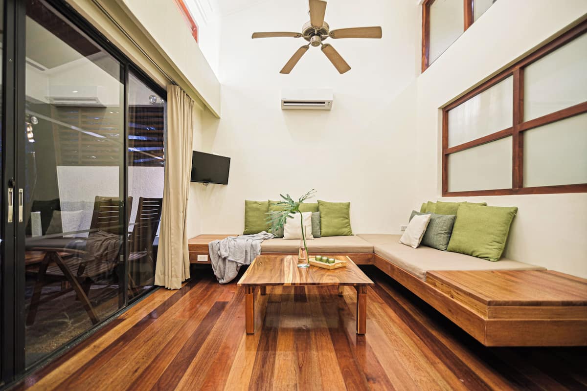 A modern living room with a wooden coffee table, a sofa with green cushions, a ceiling fan, and a TV mounted on the wall.