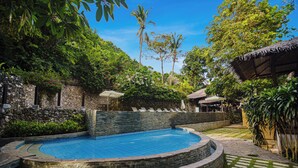 Una piscina circular con un muro de piedra, rodeada de abundante vegetación y un sendero de piedra.