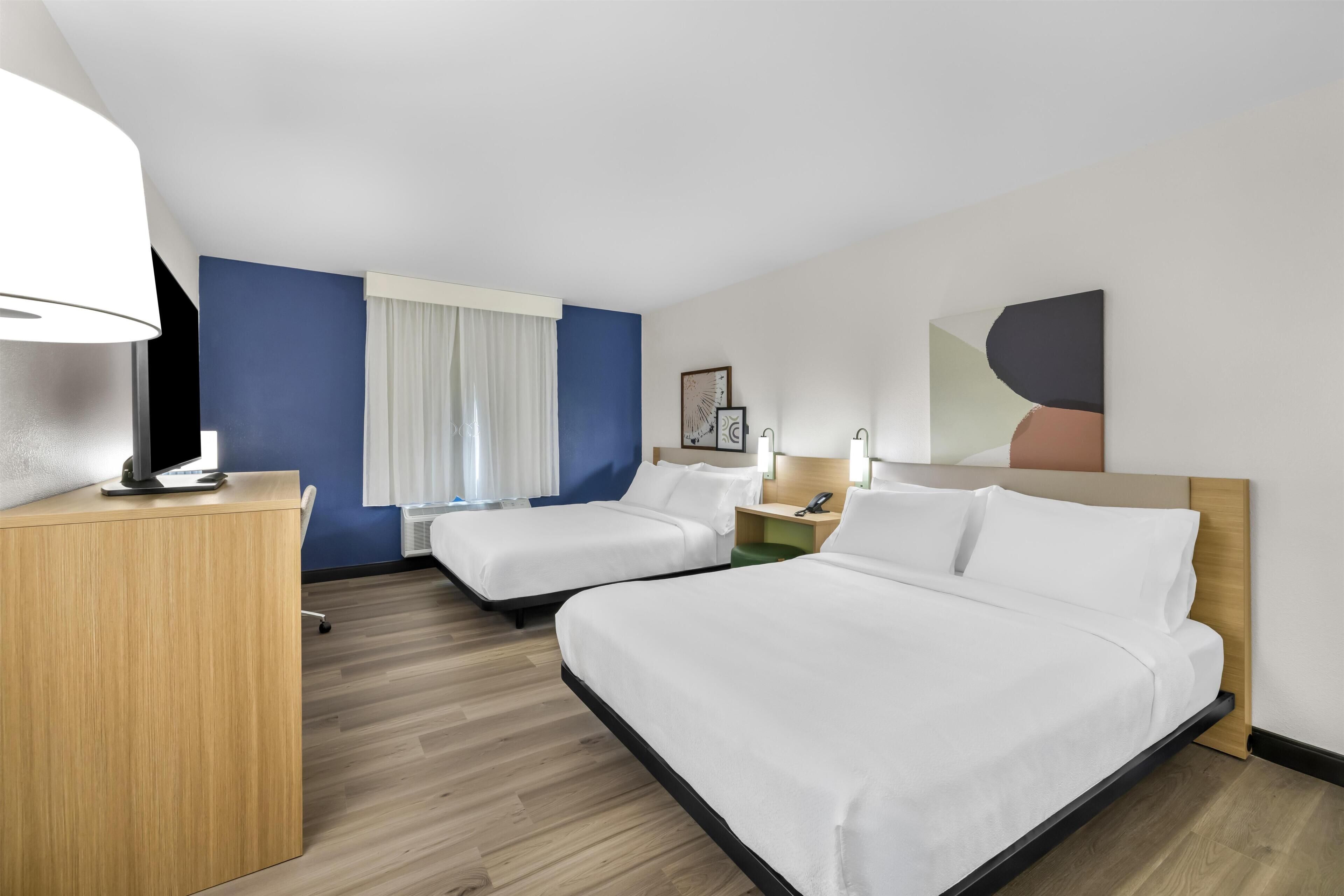 Een hotelkamer met twee bedden, een bureau, een lamp, een raam met gordijnen en kunst aan de muur.