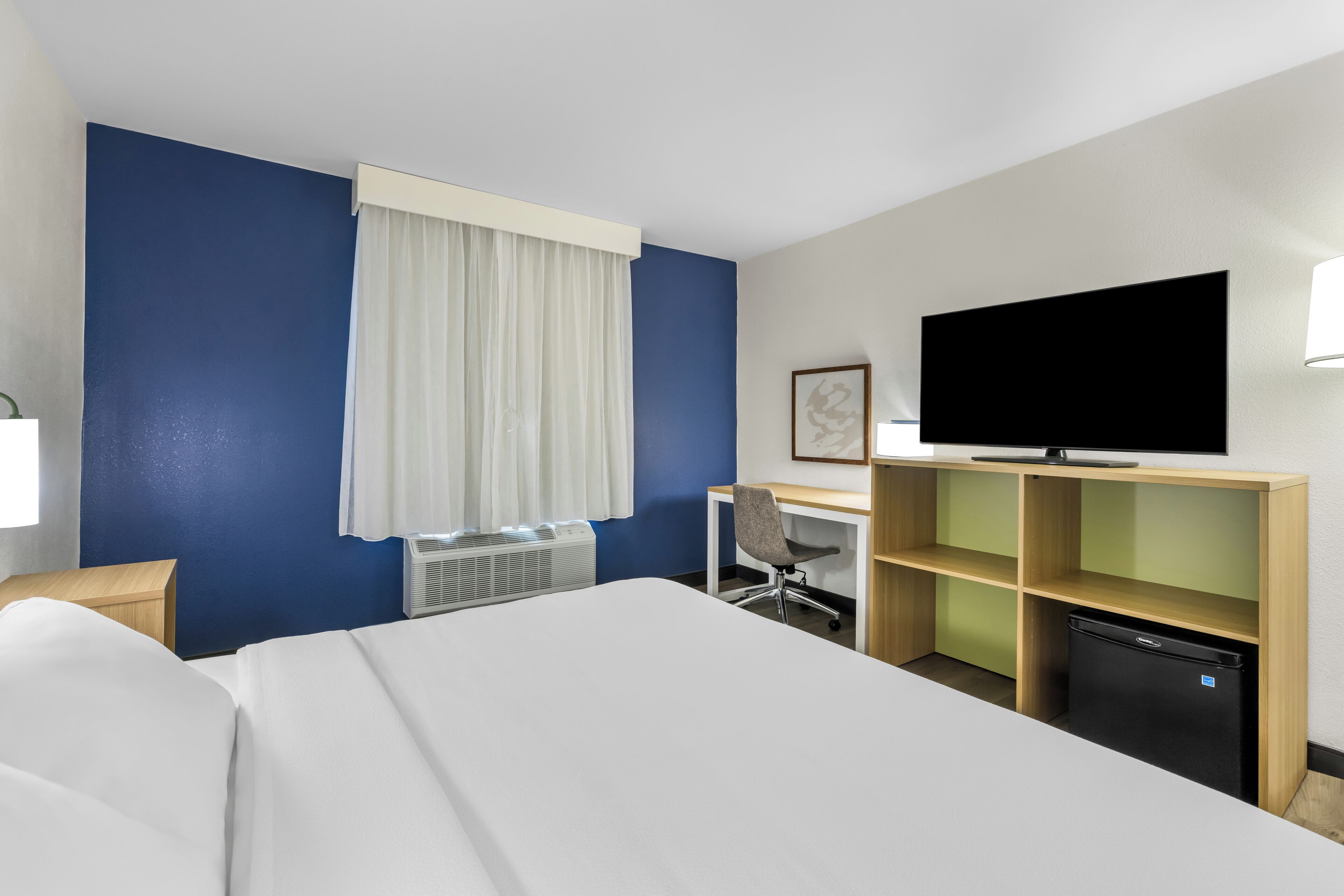 A hotel room with a bed, a desk, a chair, a television, a mini-fridge, and a window with curtains.