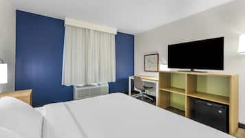 A hotel room with a bed, a desk, a chair, a television, a mini-fridge, and a window with curtains.