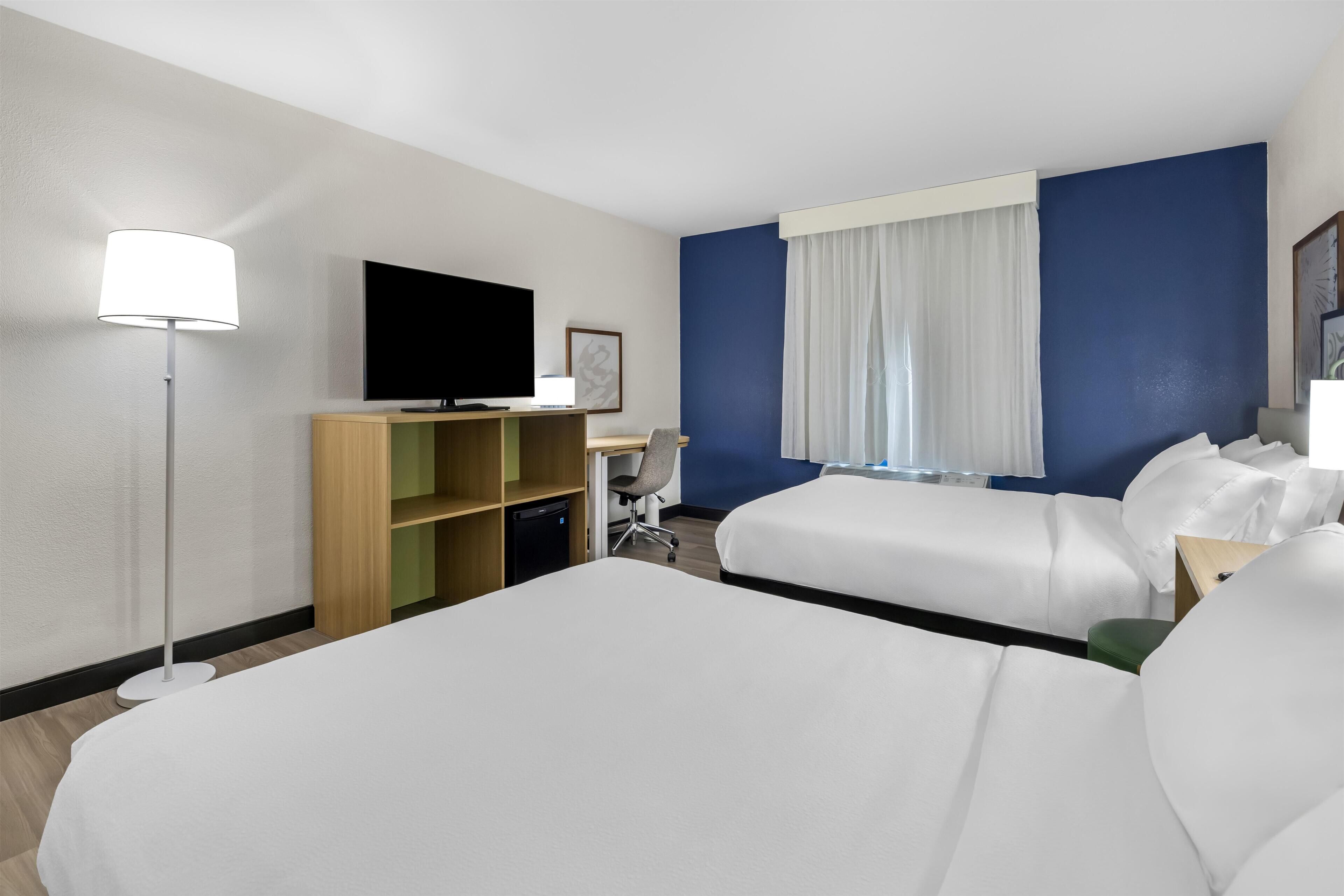 A hotel room with two beds, a desk with a computer, a TV, and a bookshelf.