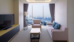 A modern hotel room with a large flat-screen TV, a sofa, a coffee table, and a view of a cityscape through large windows. - Harbor Park Hotel (Incheon)