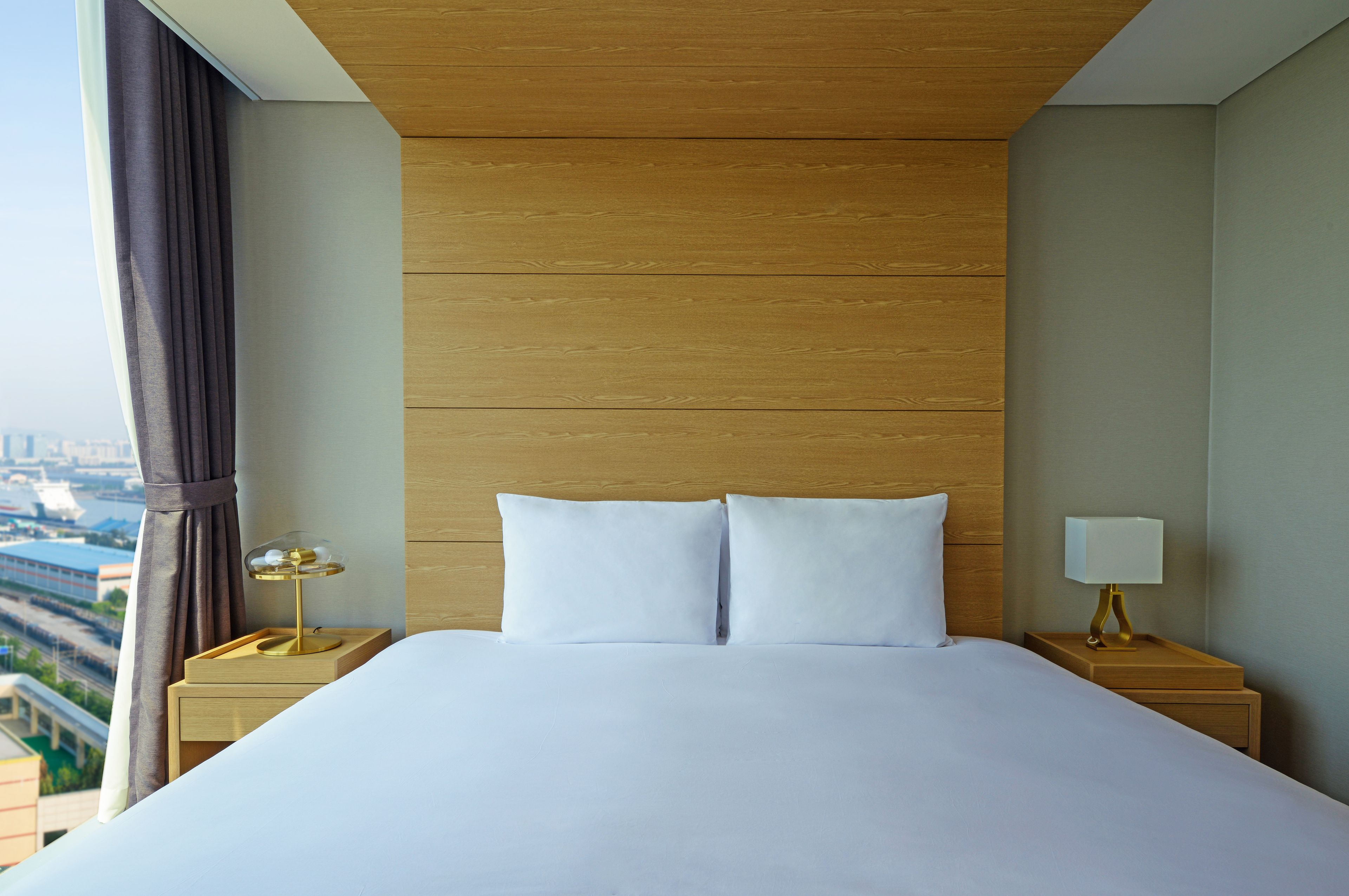 A hotel room with a large bed, two bedside tables, a lamp, and a view of a cityscape.
