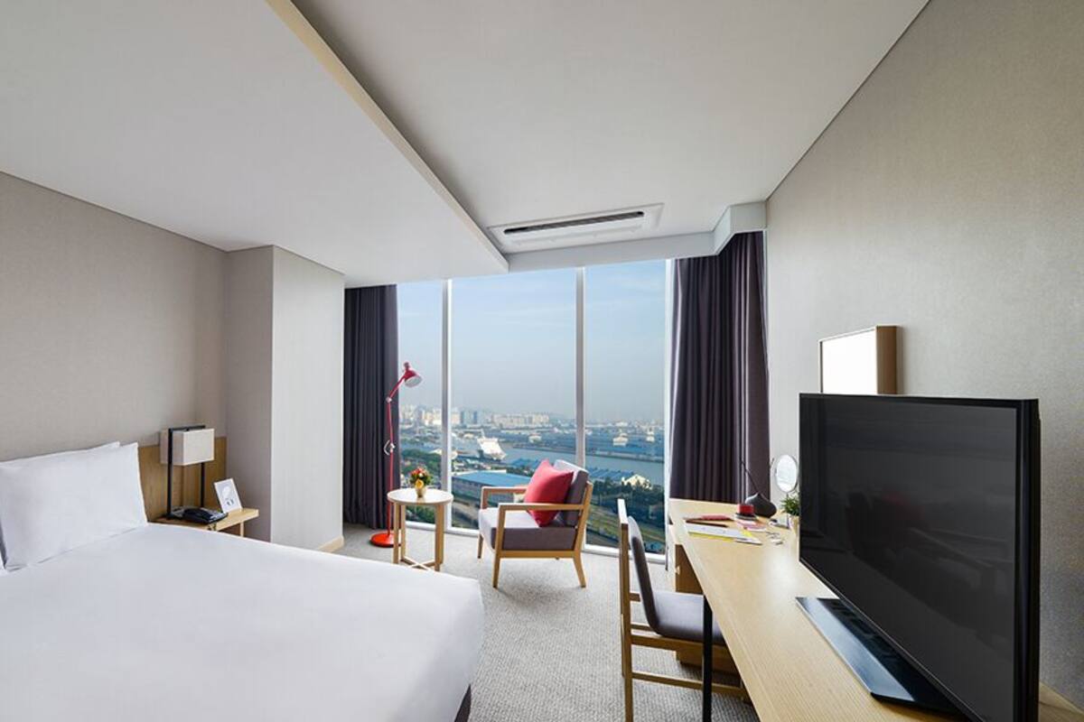 A hotel room with a large bed, a desk with a TV, a chair, a small table with a lamp, and a view of a cityscape through large windows.