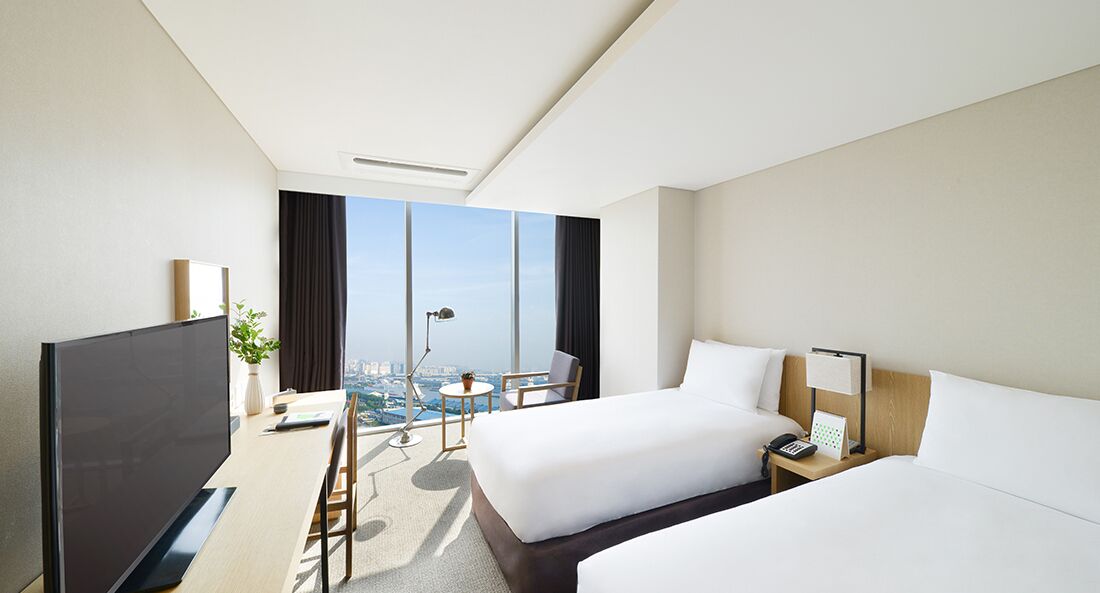 A modern hotel room with a large bed, a smaller bed, a desk, and a TV. There is a balcony with a view of the city.