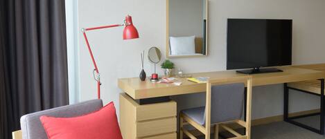 Habitación de hotel con un sofá gris, un cojín rojo, un escritorio de madera, una lámpara de escritorio roja, un televisor de pantalla plana, un espejo pequeño, una planta en maceta y una mesa redonda de madera con flores.