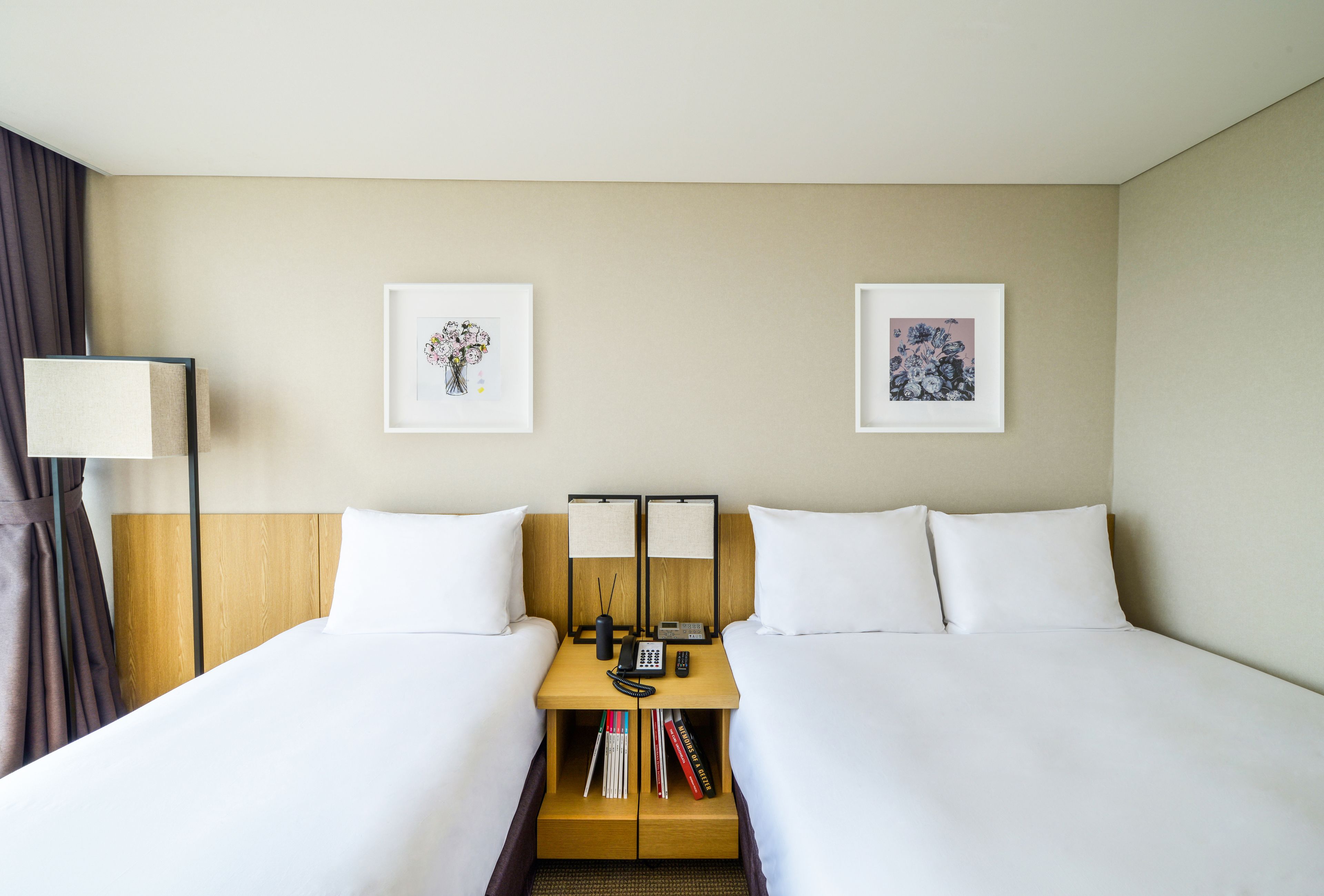 Habitación de hotel con dos camas, una mesita de noche con lámpara, teléfono y dos cuadros en la pared.