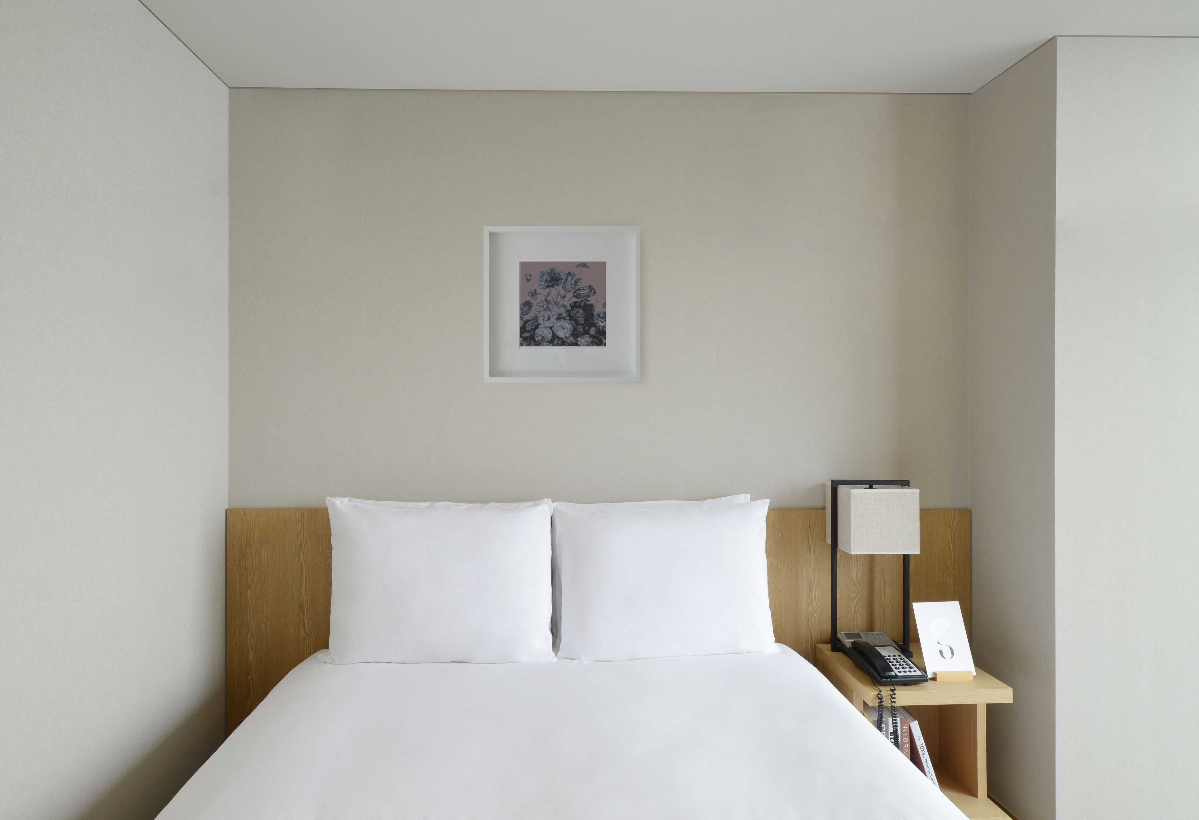 Una cama bien hecha con sábanas blancas, un cabecero de madera, una mesita de noche con una lámpara y un cuadro en la pared.
