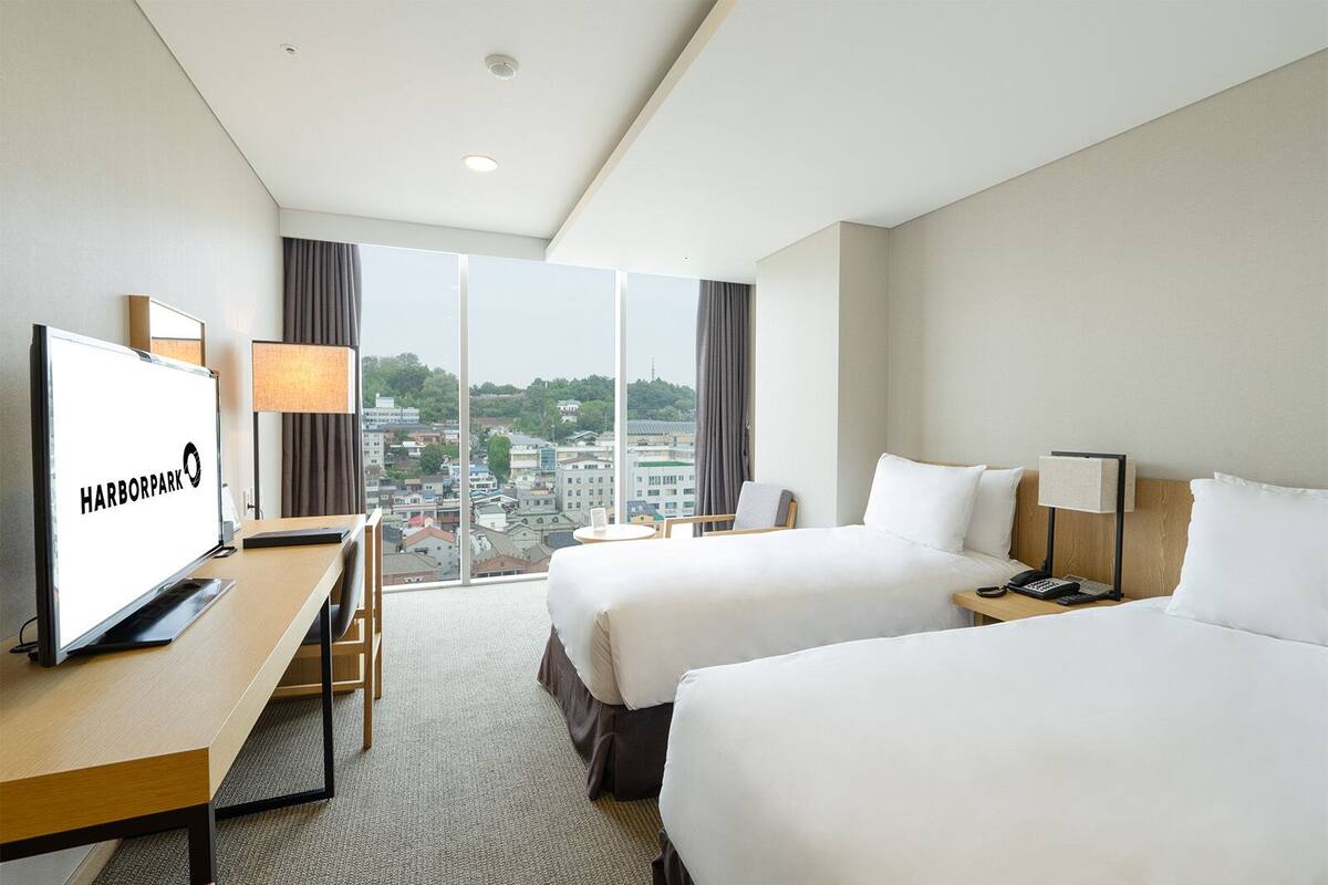 A hotel room with two beds, a desk, a TV, and a view of the city.