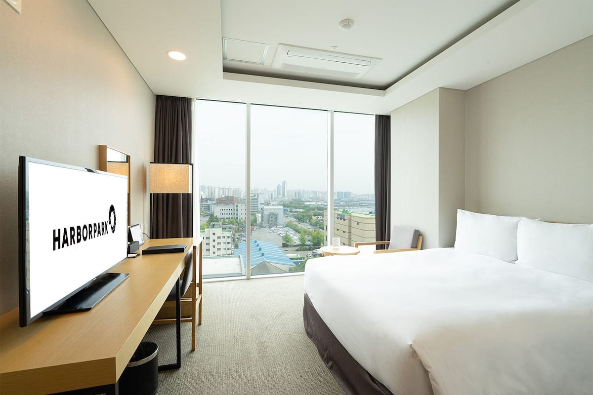 A hotel room with a large bed, a desk with a TV displaying "HARBORPARK", a city view through large windows, and a modern ceiling design.