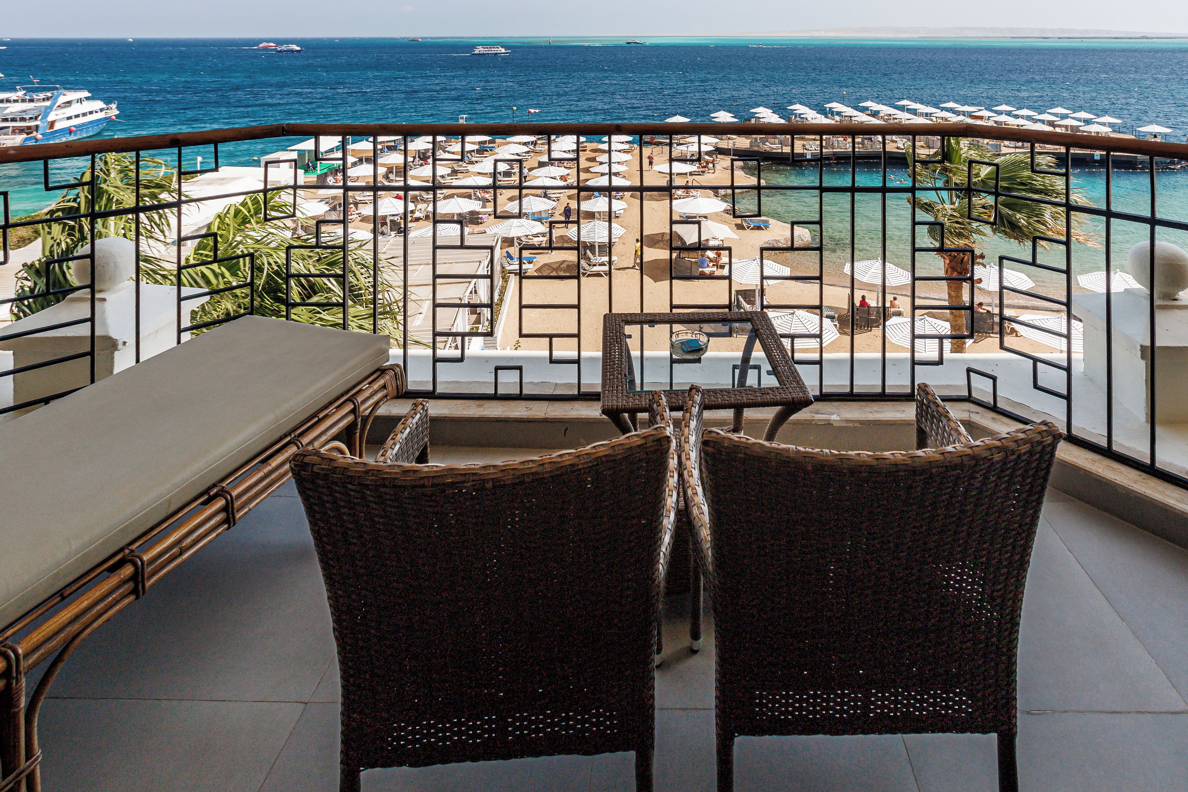 Un balcón con sillas de mimbre y una mesa, con vistas a una piscina y zona de playa con sombrillas y sillones de descanso.