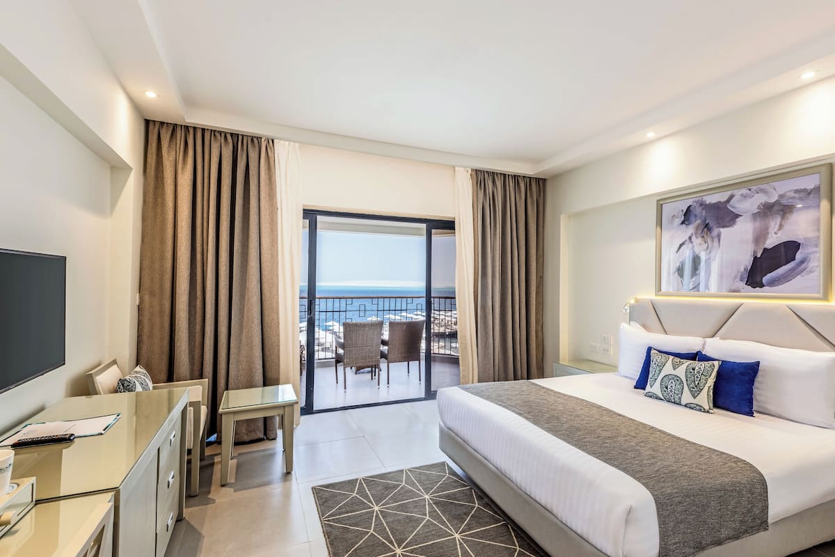 Quarto de hotel com cama, escrivaninha, televisão e varanda com vista para o mar.