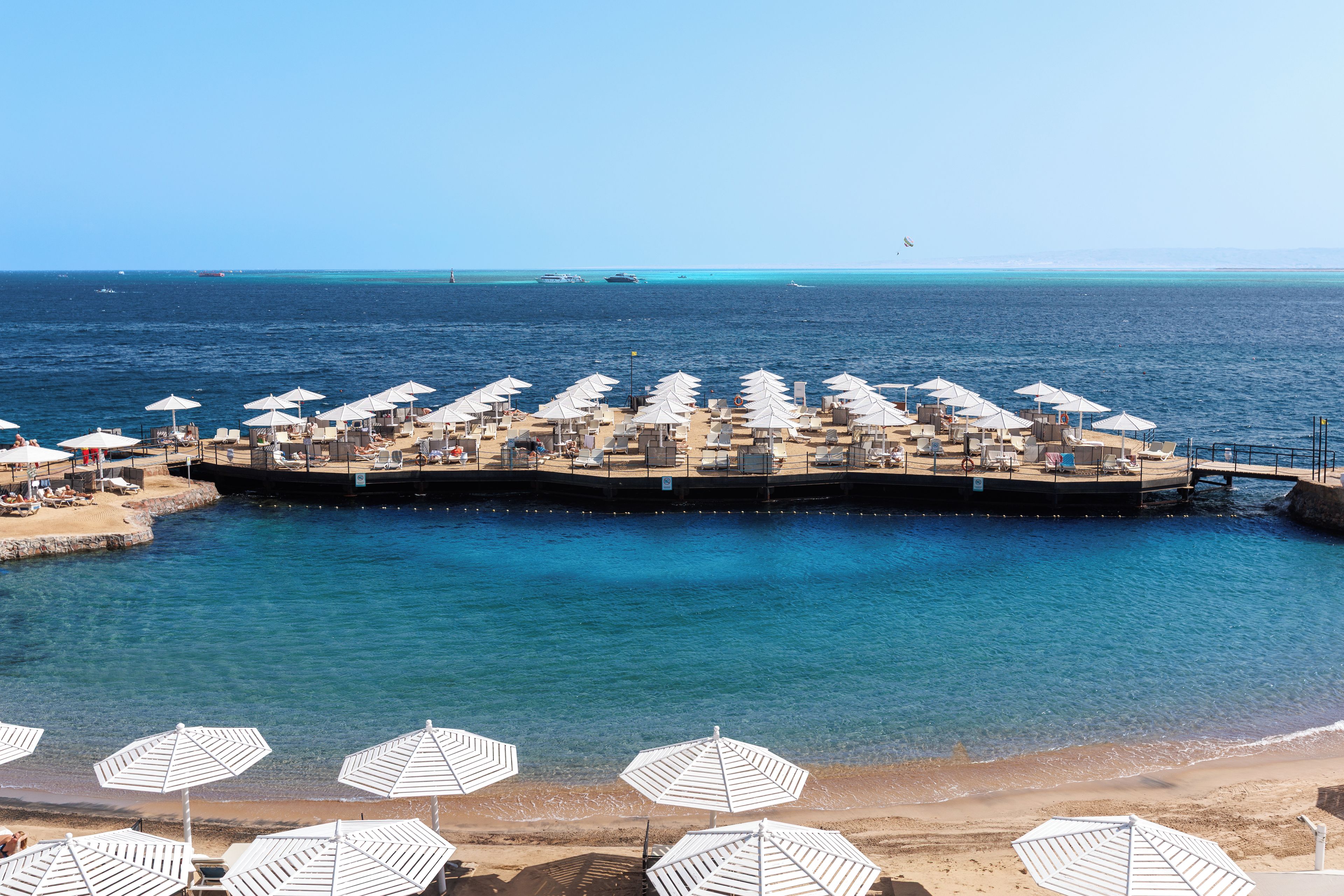 Ein Strandresort mit mehreren Liegestühlen und Sonnenschirmen, einer Pier, die ins Meer ragt, und klarem, blauem Wasser.