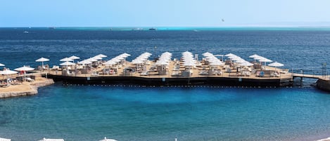 Un resort frente a la playa con múltiples tumbonas y sombrillas, un muelle que se adentra en el mar y aguas azules cristalinas.