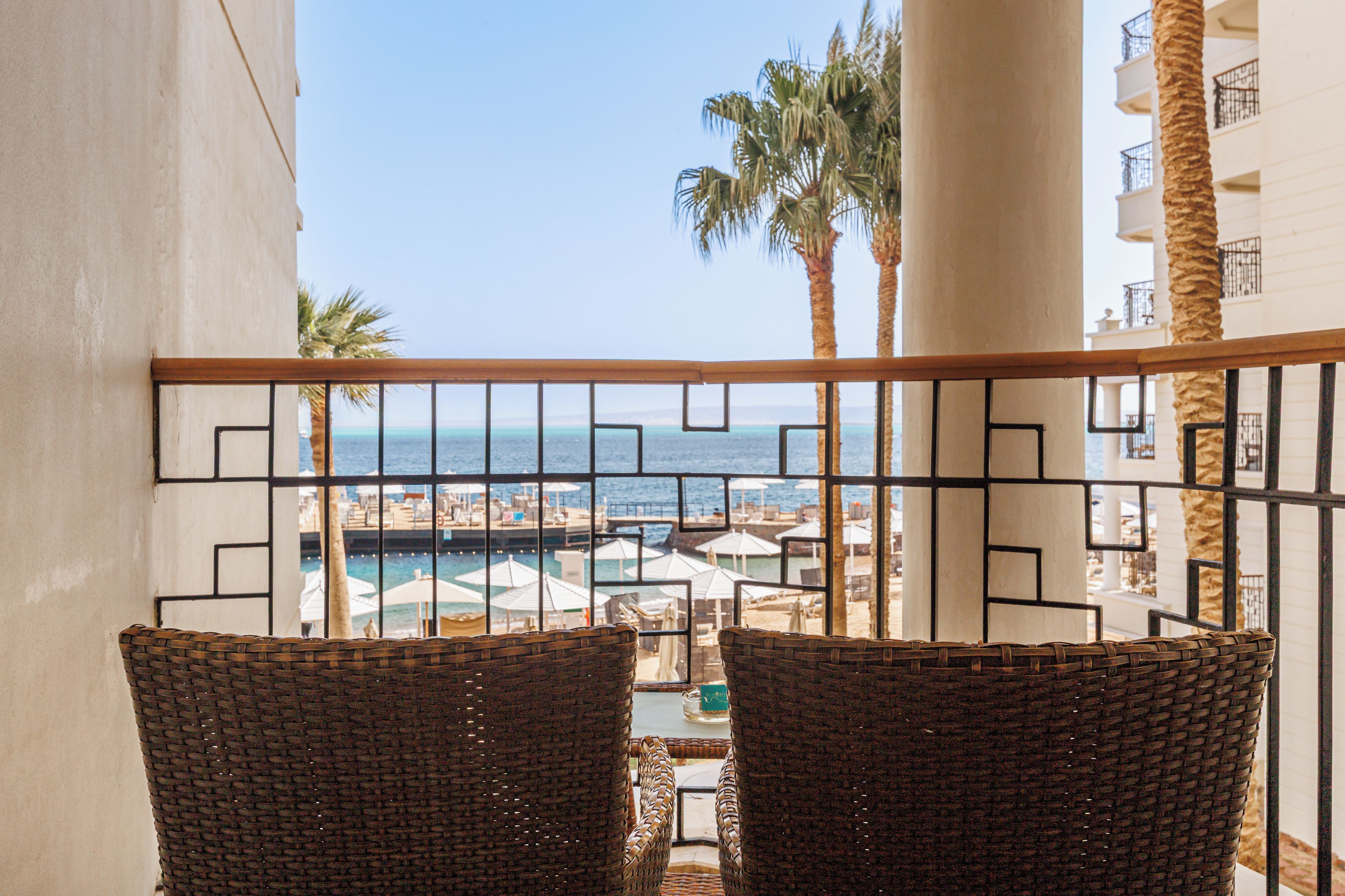 Un balcon avec des fauteuils en osier donnant sur une vue de la plage, des palmiers et une zone avec piscine.