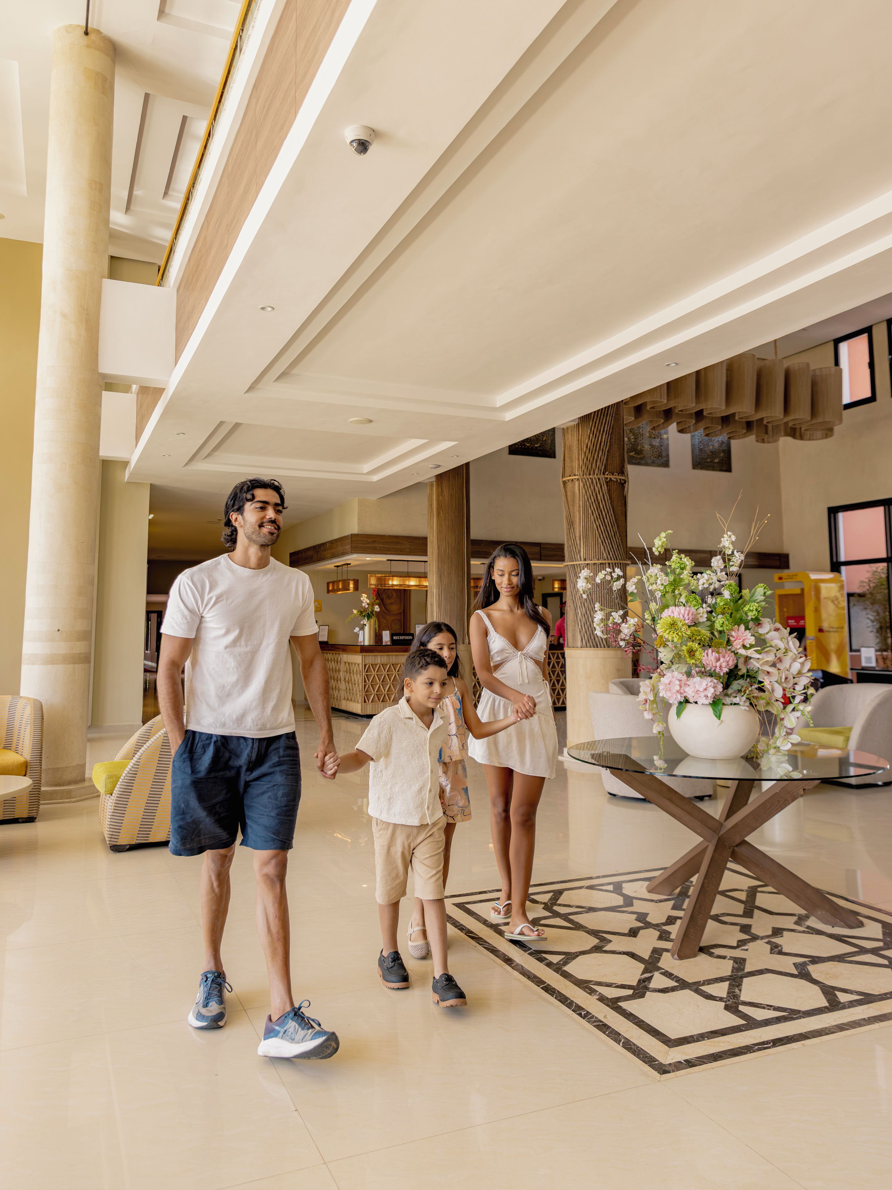 A family in a hotel lobby with a reception desk, seating area, and a decorative vase with flowers.