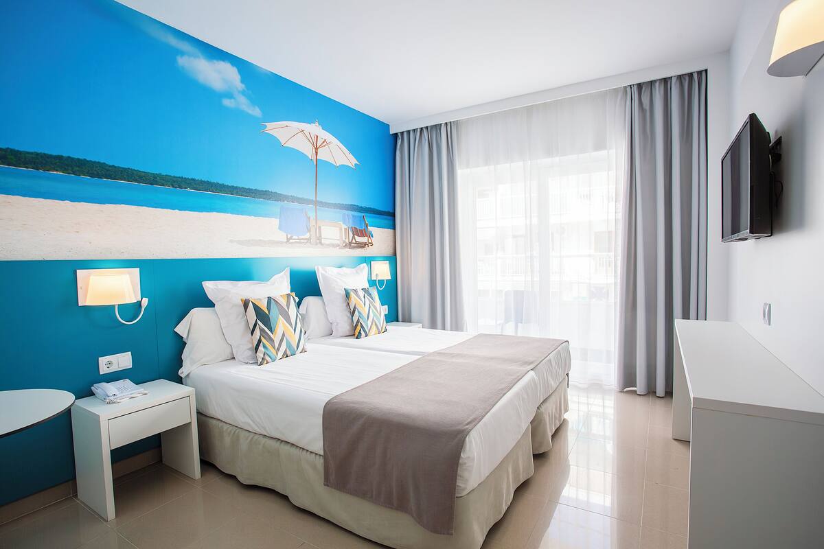 Une chambre d’hôtel avec un grand lit, une table de chevet avec un téléphone, un téléviseur à écran plat et un tableau représentant une scène de plage accroché au mur.