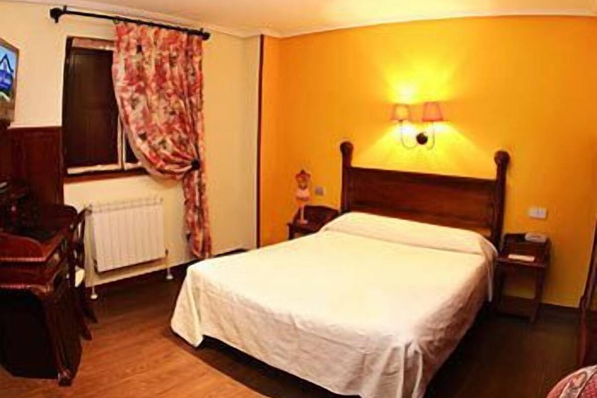A hotel room with a bed, a nightstand, a lamp, a window with curtains, and a radiator.