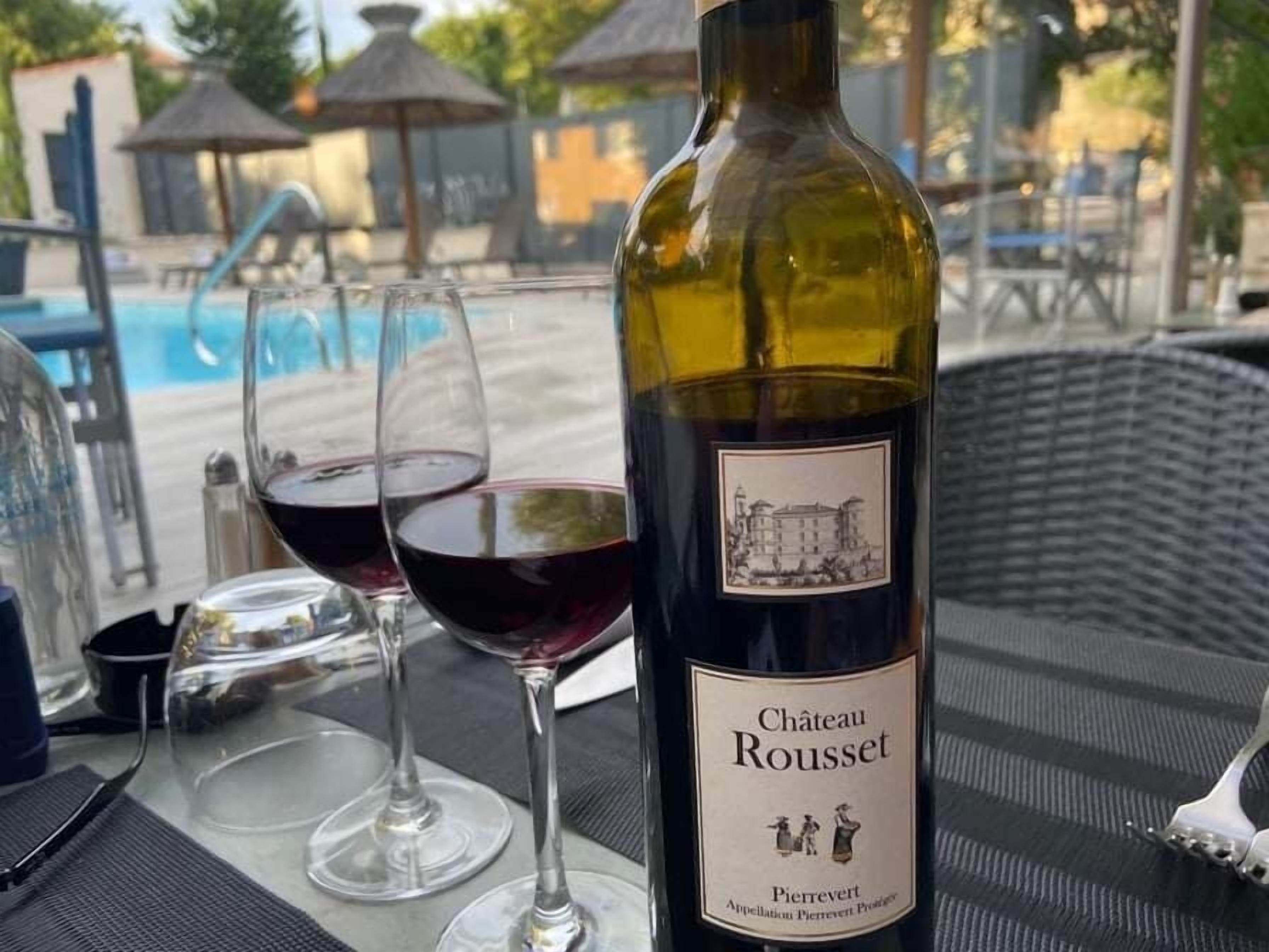 A bottle of Chateau Rousset wine with two glasses of red wine on a table near a pool area.